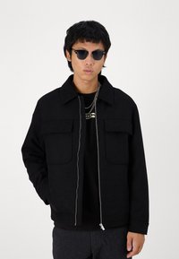 Casaco preto de fecho com gola, duas pockets frontais e uma textura suave. Usado sobre uma t-shirt preta, combinado com óculos de sol escuros.