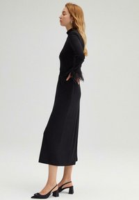 Sort kjole med lange ærmer, tætsiddende liv, frynse detaljer på ærmerne og en svajende nederdel; parret med sorte slingback hæle.