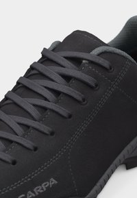 Gros plan sur une chaussure de sport en cuir noir avec des lacets gris foncé et des coutures visibles, affichant le nom de la marque "Scarpa" sur le côté.