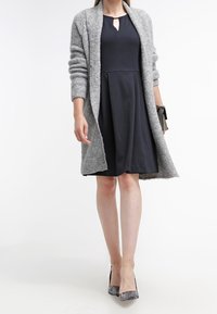 Cardigan de punto gris sobre un vestido azul marino oscuro con escote tipo ojal. Tacones grises con un patrón texturizado completan el conjunto.
