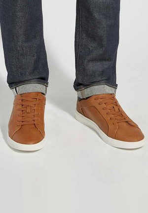 Bruine leren sneakers met witte rubberen zolen. Voorzien van platte veters en subtiele stiksels. Draagt men met donkerblauwe spijkerbroek, omgeslagen bij de enkels.