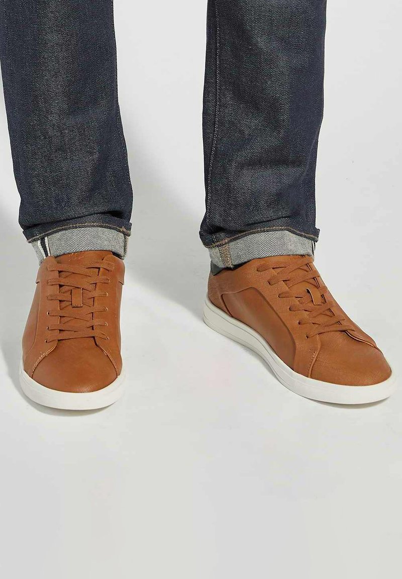 Baskets en cuir marron avec semelles en caoutchouc blanc. Elles présentent des lacets plats et des coutures discrètes. Portées avec un jean denim foncé, retroussé aux chevilles.