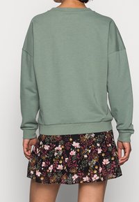 Felpa verde con vestibilità rilassata, polsini e orlo a costine. Abbinata a una gonna floreale nera con fiori rosa e bianchi.