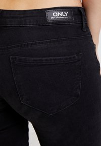 Jeans de mezclilla negros con un diseño de bolsillo trasero estándar, costuras a tono y una etiqueta de la marca en la cintura. Textura suave, forma ajustada.