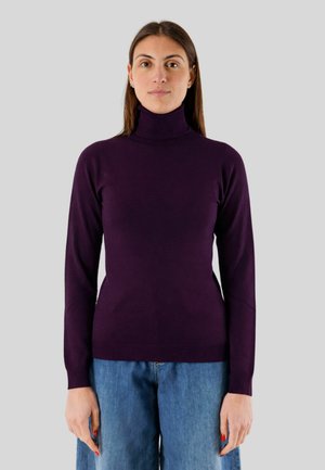 Donna con capelli castani lisci, che indossa un maglione dolcevita viola scuro e jeans blu, in piedi davanti a uno sfondo grigio uniforme.
