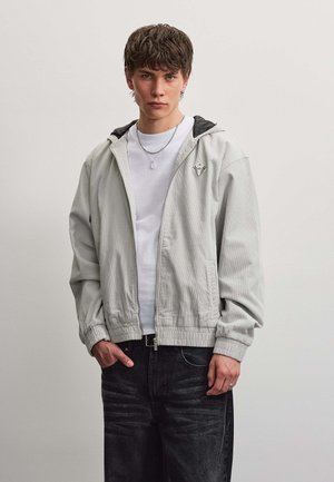 Junger Mann in hellgrauer Zip-up-Jacke und weißem Hemd steht mit einer Hand in der Tasche vor einfachem Hintergrund.