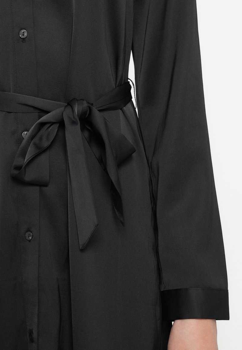 Zwarte overhemdjurk van soepel materiaal, met een gestrikt tailleband, lange mouwen en een strakke, minimalistische uitstraling met een net geplooide manchet.