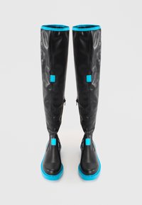 Black knee-high boots made of glossy material, featuring turquoise accents, square patches, and side zippers. Thick turquoise soles.
