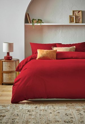 Bett mit roter Bettwäsche und zwei beigen Kissen, hölzerner Nachttisch mit roter Lampe, Regal mit Büchern, Pflanzen und gerahmter Kunst an strukturierter Wand.