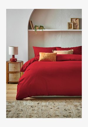 Bett mit roter Bettwäsche und zwei beigen Kissen, hölzerner Nachttisch mit roter Lampe, Regal mit Büchern, Pflanzen und gerahmter Kunst an strukturierter Wand.