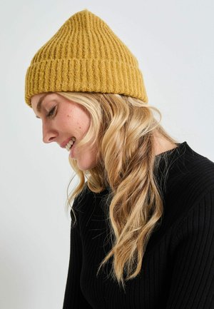 Gorro de punto acanalado amarillo mostaza con la parte superior puntiaguda, que presenta un patrón texturizado. Llevado con un suéter negro acanalado.
