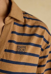 Pruuniribiline polo-särk tumesiniste triipudega, millel on krae ja vasakul rinnal tikitud "JOULES 1899" logo. Pehme tekstuur.