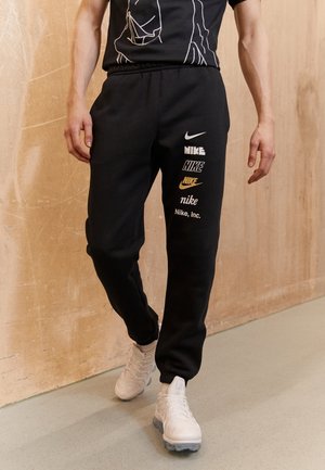 Mężczyzna w czarnych spodniach dresowych z kilkoma białymi i złotymi logo Nike na jednej nodze, białe tenisówki, spacerujący wewnątrz przy beżowej ścianie.