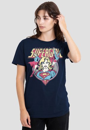 Woman wearing a navy blue T-shirt with vintage-style Supergirl comic illustrations and text, standing against a plain light background.