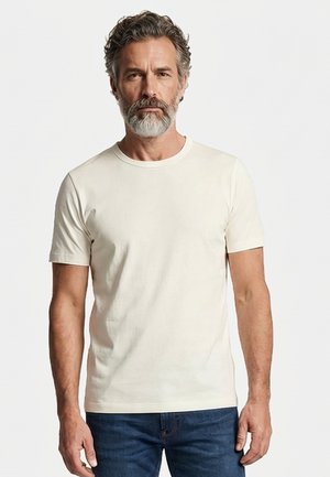 Man met grijs haar en baard, gekleed in een eenvoudige witte T-shirt met korte mouwen en blauwe spijkerbroek, staand tegen een effen lichte achtergrond.