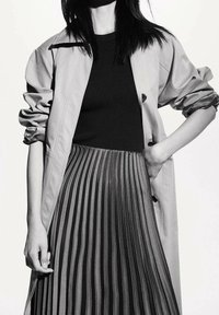 Abrigo trench beige con mangas remangadas, top negro de canalé y falda negra plisada. Los pliegues crean un aspecto texturizado y fluido.