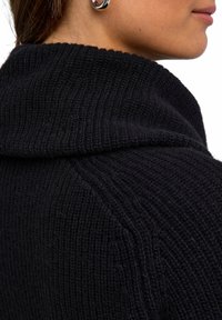 Maglione nero lavorato a maglia con texture a coste e colletto ripiegato, che mostra un primo piano della zona spalle e parte del collo.