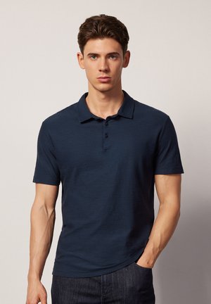 Navy-blaues Poloshirt mit Kragen, kurzen Ärmeln, Drei-Knopf-Leiste und strukturiertem Stoff, kombiniert mit dunklen Jeans.
