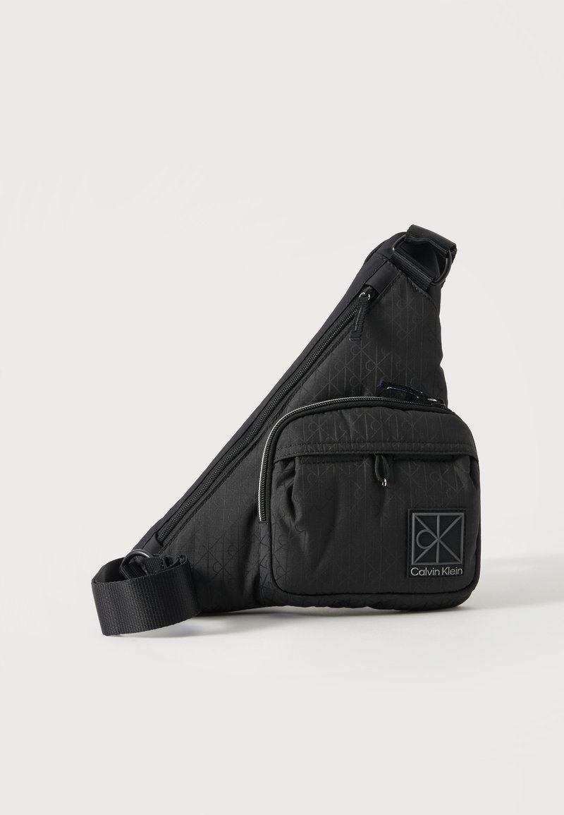 Μαύρη τσάντα Calvin Klein crossbody με ρυθμιζόμενο λουρί, μπροστινή τσέπη με φερμουάρ και διακριτικό μοτίβο λογότυπου σε απλό φόντο.