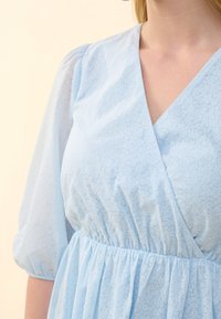 Vaaleansininen mekko, jossa on teksturoitu kukkakuosi, V-pääntie ja kiristetty vyötärö. Mekossa on lyhyet puffihihat ja liehuva kangas.