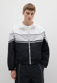 Zwart-wit jack met ritssluiting, voorzien van een hoge kraag, elastische manchetten en contrasterende strepen over de borst. Lichtgewicht materiaal.