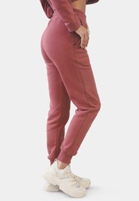 Bordeauxfarbene Jogginghose mit Seitentaschen, schmal zulaufendem Bein und elastischem Bund, kombiniert mit weißen, klobigen Turnschuhen. Sanfte Textur.