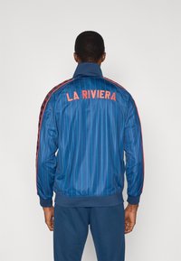 Μπλε ριγωτό τζάκετ με φερμουάρ, με κεντημένο το "LA RIVIERA" σε πορτοκαλί στο πίσω μέρος και κόκκινα πλαϊνά διακοσμητικά, κατασκευασμένο από ένα λείο υλικό.