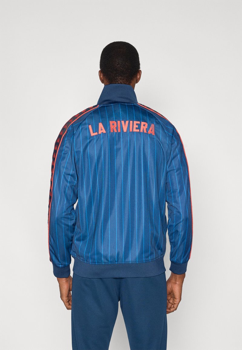 Μπλε ριγωτό τζάκετ με φερμουάρ, με κεντημένο το "LA RIVIERA" σε πορτοκαλί στο πίσω μέρος και κόκκινα πλαϊνά διακοσμητικά, κατασκευασμένο από ένα λείο υλικό.