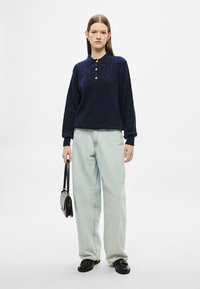 Μπλε σκούρο πλεκτό πουλόβερ με γιακά και τρία χρυσά κουμπιά, συνδυασμένο με ανοιχτό γαλάζιο παντελόνι φαρδιάς γραμμής και μαύρα loafers, κρατώντας μια μαύρη τσάντα.