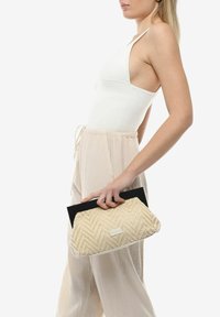 Clutch tejido beige con un patrón de espiga, detalle negro y ribete blanco. Sostenido en una mano mientras se veste ropa de color claro.