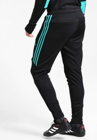 Person in schwarzen Sporthosen mit türkisfarbenen Streifen und schwarzen Adidas Copa-Schuhen, posiert mit einem leicht angehobenen Fuß.