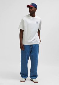 Hvid T-shirt med korte ærmer og logo, kombineret med løse blå denimjeans og hvide sneakers med grønne detaljer. Rød og blå baseballkasket.