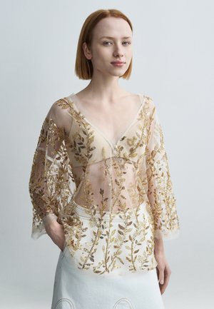 Gennemsigtig langærmet top med guldbroderi i form af blade, V-hals design og gennemsigtigt stof over et hvidt skørt med blomsterdetaljer.