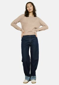 Maglione beige a maglia strutturata con scollo rotondo. Jeans in denim blu scuro a gamba larga con risvolto a contrasto. Modella in piedi, mani sui fianchi.