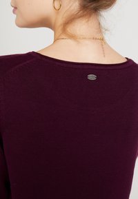 Burgundy neulepaita, jossa on hieno kudontatekstuuri. Ominaisuuksiin kuuluu pyöreä pääntie ja pieni hopeinen logotagi takana, lähellä kaulaa.