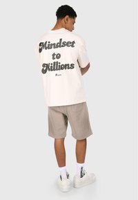 Cremefarvet bomulds t-shirt med stor sort tekst på ryggen, der står "Mindset to Millions", parret med beige shorts og hvide sneakers.