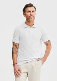 Biała koszulka polo z klasycznym kołnierzykiem, krótki rękaw i zapięcie na guziki. Noszona z beżowymi spodniami; widoczne tatuaże na ramieniu.