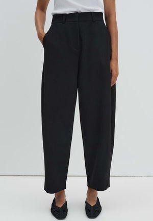 Femme portant un pantalon noir taille haute à jambes larges avec poches latérales, associé à des ballerines noires pointues et un top blanc sans manches.