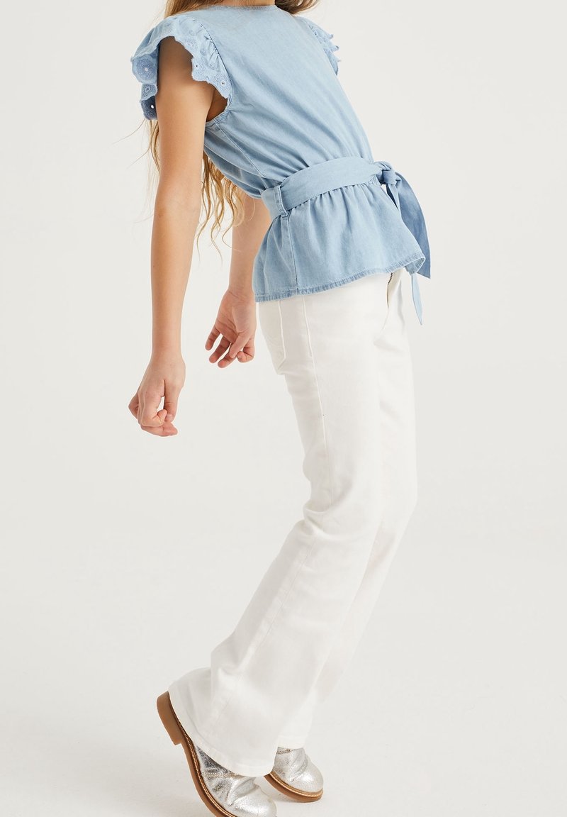 Fille portant un chemisier bleu clair ceinturé, un pantalon blanc et des chaussures argentées, penchée en arrière contre un fond blanc uni.