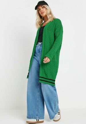 Cardigan vert en tricot avec un design texturé, ouverture à l'avant, manches longues et accents rayés noirs aux poignets. Associé à un jean à jambes larges.