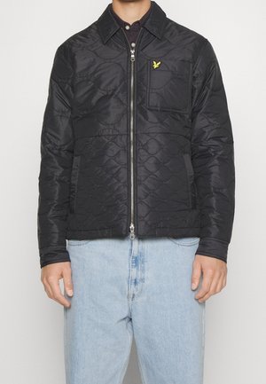 Hombre vestido con una chaqueta acolchada negra con cremallera y un logo de pájaro bordado amarillo en el bolsillo del pecho, junto con unos jeans de color azul claro.