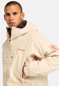 Beige Timberland-takki, jossa on huppu, oranssit yksityiskohdat, vetoketjusuljenta ja elastiset hihansuut. Teksturoitu kangas etutaskuilla ja logoyksityiskohdalla.