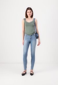 Débardeur vert avec des détails en dentelle, associé à un jean skinny bleu clair et des chaussures plates noires. Modèle tenant un sac à bandoulière bleu. Fond neutre.