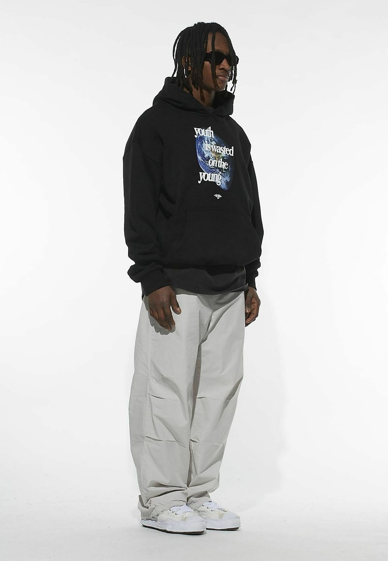 Sudadera negra con texto blanco y un gráfico azul, combinada con pantalones oversize gris claro y zapatillas blancas. Modelo con gafas de sol oscuras.