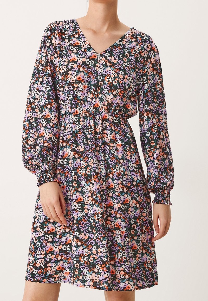 Kukallinen mekko mustalla pohjalla ja eläville väreille; siinä on V-pääntie, pitkät puffihihat ja tyköistuva vyötärö. Pehmeä kangas, jossa on virtaava muotoilu.