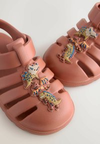 Ružové gumené sandále s dinosaurími dekoráciami, vybavené nastaviteľným popruhom, otvoreným dizajnom a ventilačnými otvormi pre priedušnosť.
