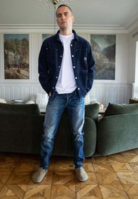 Chaqueta de mezclilla de color azul oscuro, camiseta blanca por debajo, jeans de color azul claro y zapatos tipo slip-on de color gris claro, de pie en una habitación con suelo de madera.