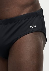 Κοντινό πλάνο ενός ατόμου που φοράει μαύρο μαγιό με τη λέξη «BOSS» τυπωμένη λευκή στο πλάι.