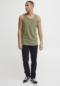 Olivgrünes Tanktop mit Rundhalsausschnitt und Brusttasche, kombiniert mit schwarzen Slim-Fit-Hosen und schwarzen Sneakers mit weißen Akzenten.