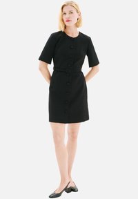 Robe noire à manches courtes avec un col rond, des boutons sur le devant et une taille ceinturée. Texture de tissu lisse, design à longueur genou, associée à des chaussures noires.